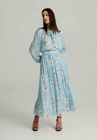Light blue maxi dress with floral print, long sleeves, a tie neck, and gathered waist. Paired with black heeled sandals. Smooth fabric.