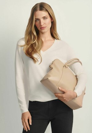 Kobieta z długimi blond włosami, ubrana w biały sweter i czarne spodnie, trzymająca dużą beżową torebkę na jednolitym tle.