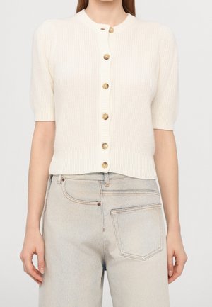 Kvinde iført en cremefarvet kortærmet strikket cardigan med seks brune knapper, kombineret med lysegrå højttaljede jeans.