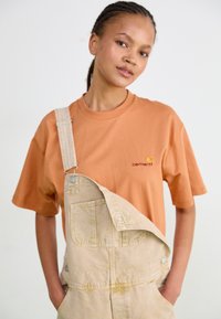 T-shirt orange avec le logo Carhartt, associé à des salopettes beige clair dotées d'une poche et de bretelles ajustables. Coupe décontractée et confortable.