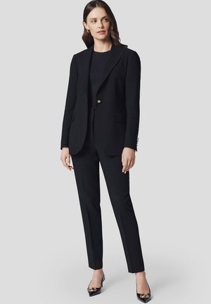 Czarny dopasowany garnitur z jednorzędową marynarką z klapkami i slim-fit spodniami. W połączeniu z czarnymi spiczastymi szpilkami.