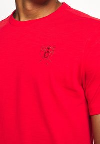 Hombre de camiseta de cuello redondo roja brillante con un pequeño logotipo bordado de un pantera rugiente en el lado izquierdo del pecho.