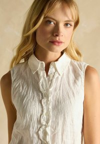 Bluză din in albă fără mâneci, cu un plastron cu volane, textură moale și guler clasic. Design minimalist, fără modele.