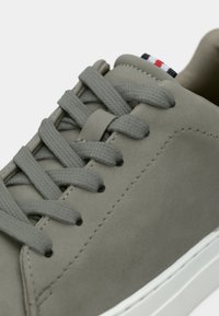 Baskets en daim gris avec lacets plats gris, semelle blanche et coutures simples. Petit détail de logo en rouge, blanc et bleu sur le talon.