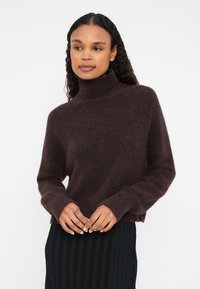 Jersey de cuello alto burdeos, hecho de un material suave y texturizado, con mangas largas y un corte holgado, combinado con una falda negra plisada.