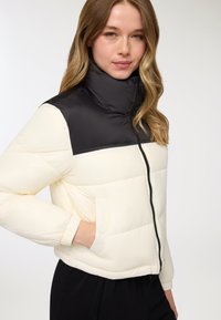 Giacca imbottita con parte superiore nera e sezione inferiore crema. Presenta un colletto alto, zip frontale e polsini elasticizzati. Texture liscia e trapuntata.