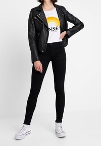 Chaqueta de cuero negra de motorista, camiseta blanca con gráfico de un atardecer en amarillo y naranja, jeans ajustados negros y zapatillas blancas con rayas negras.
