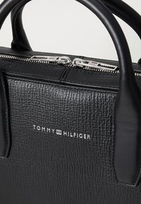 Sort læder tote taske med struktureret overflade, sølvhardware og lynlåslukning. Har "TOMMY HILFIGER" logoet i hvid.