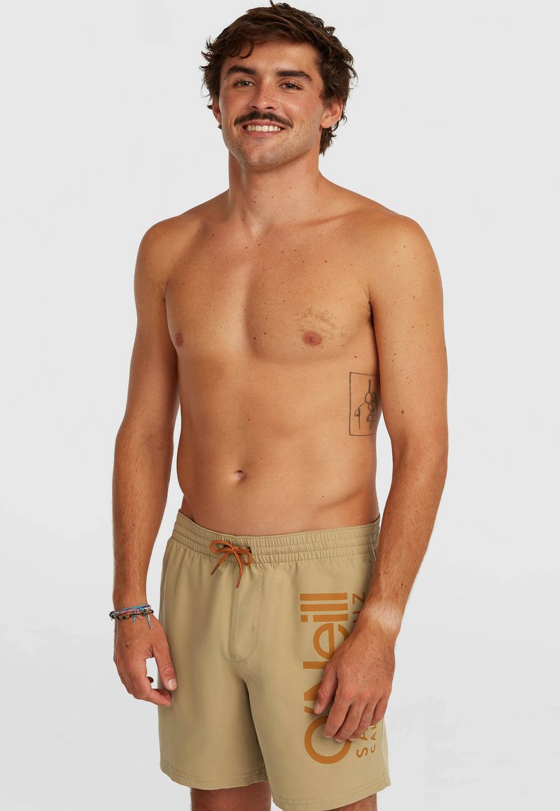 Smilende mand uden skjorte med tatovering på venstre ribben iført beige badeshorts med orange snor og side-logo, stående mod en ensfarvet baggrund.