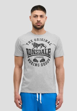 Mand iført grå Lonsdale London boksning T-shirt og blå shorts med snor, stående mod en ensartet lys baggrund.