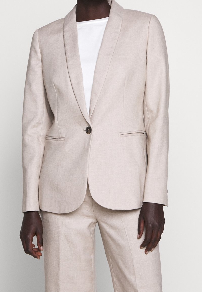 Personne à la peau foncée portant un blazer beige clair ajusté avec un bouton et un pantalon assorti, accompagné d'un haut blanc.
