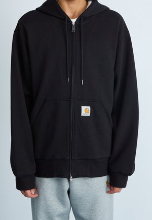 Person trägt einen schwarzen Kapuzenpullover mit Reißverschluss, aufgesetzter Tasche vorne und Carhartt-Logo-Patch sowie hellgraue Jogginghosen.