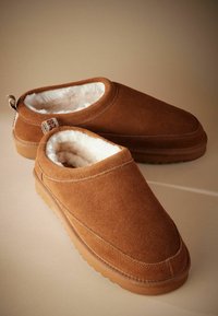 Brune ruskinds slip-on sko med en luksuriøs hvid pelsforing, en rund tå og en tekstureret gummisål med en bageste trækketag.