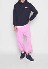 Persona vestida con una sudadera negra con capucha, pantalones rosa claro y zapatillas azul marino con el logotipo de Ewing en amarillo y rojo, de pie con una mano en la cadera.