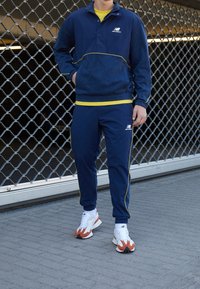 Námořnicky modrá tepláková souprava se žlutým lemem, obsahující vršek na zip a kalhoty s elastickým pasem. Nositelná s bílými a oranžovými sportovními botami.