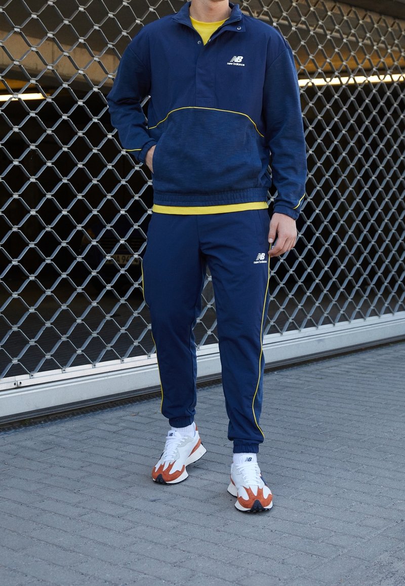 Námořnicky modrá tepláková souprava se žlutým lemem, obsahující vršek na zip a kalhoty s elastickým pasem. Nositelná s bílými a oranžovými sportovními botami.