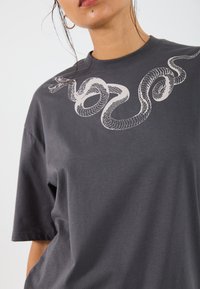 T-shirt en coton gris avec un graphique de serpent blanc le long du col, des manches courtes et une coupe décontractée. Texture lisse.
