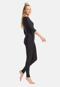 Lyshåret kvinde iført sort tætsiddende langærmet top og leggings, stående barfodet, smilende og gående foran en enkel hvid baggrund.