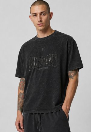 Giovane uomo con capelli corti che indossa una maglietta nera scolorita Richmond London e pantaloni neri, mostrando braccia tatuate e mani nelle tasche.