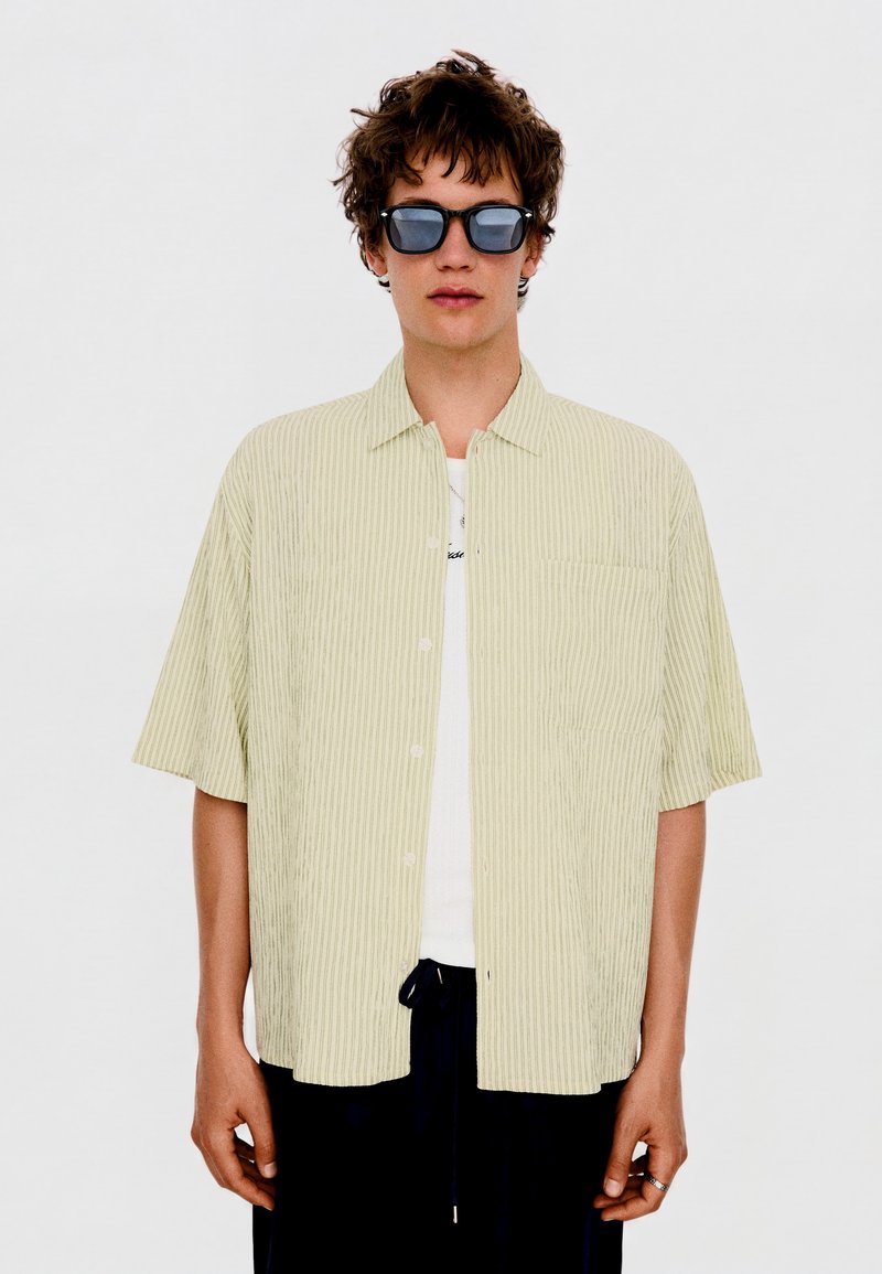 Jeune homme portant des lunettes de soleil noires, une chemise beige à rayures manches courtes boutonnée par-dessus un T-shirt blanc, et un pantalon noir sur un fond uni.