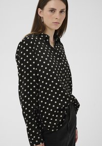 Hosszú ujjú fekete ing fehér pöttyökkel, gombos gallérral és sima szövésű anyaggal. Az ing laza szabású és ívelt alsó szegéllyel rendelkezik.