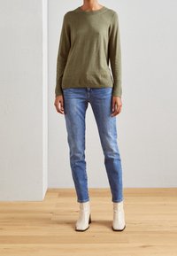 Olivgrüner Strickpullover mit langen Ärmeln, Rundhalsausschnitt, kombiniert mit hellblauen Slim-Fit-Jeans und weißen Stiefeletten. Holzboden im Hintergrund.