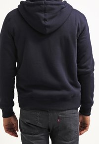 Sweat à capuche bleu marine avec capuche, poignets et ourlet côtelés, et une finition texturée. Porté avec un pantalon gris foncé, affichant un look décontracté.