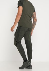 Man med mörkgrön kortärmad skjorta, matchande joggbyxor med sidofickor, svarta strumpor och mörka sportskor, står vänd bort.