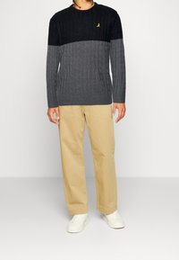 Person in einem schwarz-grau gestreiften Strickpullover, beigen Hosen und weißen Sneakers, der vor einem schlichten hellen Hintergrund steht.