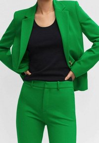 Zielona marynarka o dopasowanym kroju oraz pasujące do niej spodnie z gładkiego materiału, w połączeniu z dopasowanym czarnym topem na ramiączkach; marynarka ma jeden guzik.