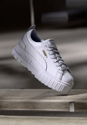 Witte Puma-sneaker met dikke geribbelde zool en veters, zwevend afgebeeld boven een betonnen oppervlak tegen een neutrale achtergrond.