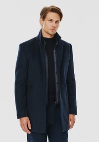 Granatowy płaszcz z wełny w składzie mieszanym, o dopasowanym kroju, z kołnierzem stojącym, kieszenią naszywaną i gładką podszewką, noszony na czarnym swetrze.