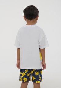 Maglietta bianca di cotone a maniche corte, abbinata a pantaloni corti blu con motivo di Pikachu giallo. Vista da dietro.