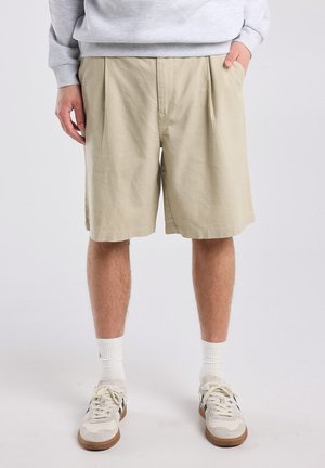 Persoon die beige knielange shorts, witte sokken en witte sneakers draagt, staand met handen in de zakken tegen een effen achtergrond.