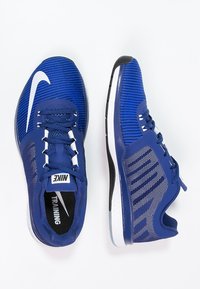 Tréninkové boty Nike z modrého síťovaného materiálu. Obsahují bílou podpisovou logo, strukturované síťované panely a černobílou podešev.