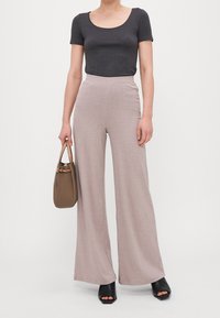 Persona con una blusa ajustada de manga corta en gris oscuro, pantalones beige de pierna ancha de cintura alta, zapatos de tacón abiertos negros, sosteniendo un bolso marrón.