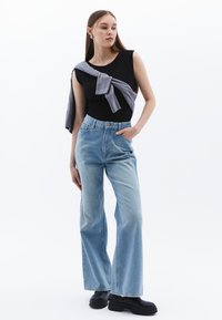 Svart ärmlös topp i kombination med ljusblå högmidjade vida jeans, kompletterad med en grå tröja knuten vid axlarna. Svarta platåskor.