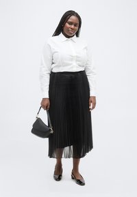 Γυναίκα με πλεγμένα μαλλιά στέκεται φορώντας λευκό πουκάμισο με κουμπιά, μαύρη midi φούστα με πιέτες, μαύρα τακούνια και κρατώντας μια μαύρη τσάντα χειρός.