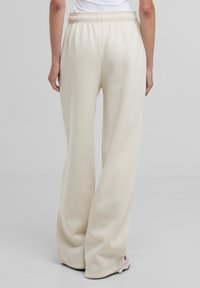 Pantaloni della tuta a gamba larga di colore crema con vita arricciata, realizzati in tessuto morbido. Caratterizzati da una texture liscia e una vestibilità rilassata.