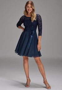 Laivansinis lyhyt suomisetti, jossa on pitsikorsetti ja kolmannen osan hihat, kelluva sifonkihame, samettinen vyönauha ja hopeiset nauhakengät.