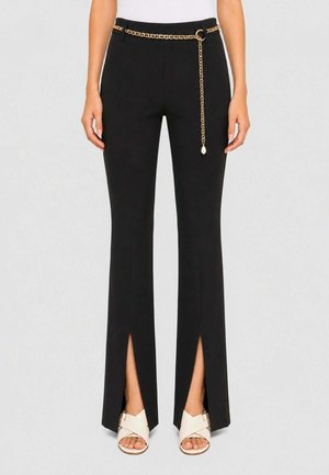 Donna che indossa pantaloni neri con spacchi anteriori sulle gambe e cintura a catena dorata, abbinati a sandali bianchi e un top bianco.