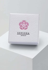 Balta teksturēta rotaslietu kaste ar rozā zieda logotipu un melnā tekstā rakstītu "SENSORA", ar kvadrātveida formu un tīru dizainu.