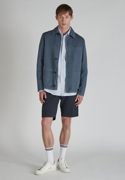 Giacca di cotone blu con chiusura a bottoni, tasca sul petto, indossata sopra una camicia bianca a righe, abbinata a pantaloni corti blu navy e sneakers bianche.