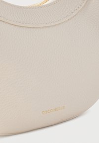 Μπεζ δερμάτινη τσάντα με στρογγυλεμένο σχήμα, υφή στην επιφάνεια, χρυσλό φερμουάρ και ανάγλυφο όνομα της μάρκας "Coccinelle" με χρυσά γράμματα.