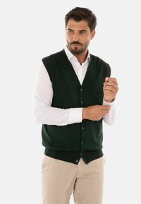 Gilet lavorato a maglia verde scuro con scollo a V, con bottoni e orlo a costine. Indossato su una camicia bianca a maniche lunghe e pantaloni beige.
