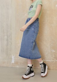 Džínová midi sukně s rozšířeným lemem, bočními kapsami a vyšívaným detailem. V kombinaci se zeleným tričkem a černými vysokými teniskami.