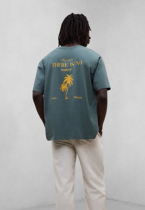 Hombre con rastas oscuras que lleva una camiseta verde azulado con texto amarillo y un gráfico de palmera, combinado con pantalones beige claros, de espaldas.