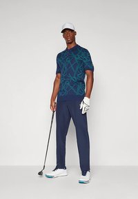 Marinblå piké med ett abstrakt mönster i teal, kombinerad med marinblå byxor. Accessoarer inkluderar en vit keps och golfhandske samt vita golfsko.