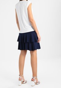 Top blanco de manga corta acompañado de una falda azul marino con volantes en capas. El modelo lleva tacones plateados con tiras. Tejido suave con un diseño en capas.