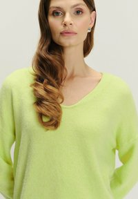 Lime zelena pletena bluza s V-izrezom i opuštenim krojem. Mekana tekstura; model ima dugu kosu stiliziranu u labavim valovima.